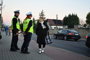 Umundurowaniu policjanci na chodniku rozmawiają ze starszą kobietą, w tle po drodze poruszają się pojazdy i cmentarz.