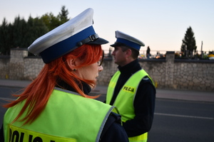 Na pierwszym planie policjanta w  umundurwaniu służbowym, w tle policjant ulica i cmentarz.