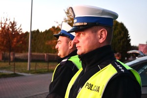 Na zdjęciu dwóch policjantów w umundurowaniu służbowym. W tle cmentarz.