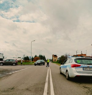 droga, obok niej radiowóz, w centralnej części policjant w umundurowaniu służbowym kierujący ruchem.