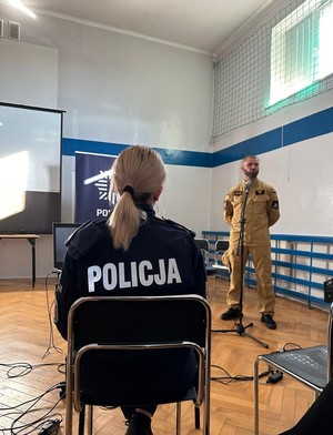 Hala gimnastyczna prelekcje prowadzi strażak w umundurowaniu służbowym, plecami siedzi policjantka w umundurowaniu służbowym.