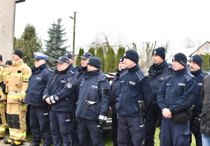 W dwuszeregu stoją policjanci w umundurowaniu służbowym, w tle domy i trawa.