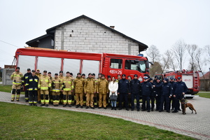 Grupa strażaków i policjantów stoją przed wozem bojowym straży pożarnej.