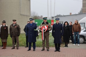 Delegacja służb mundurowych w umundurowaniu galowym z wiązanką, w tle budynki.