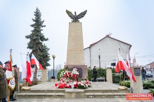 Na pierwszym planie Pomnik Niepodległości, na nim złożone kwiaty i wiązanki w tle budynki.