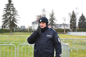 Policjant w umundurowaniu służbowym, nadaje komunikat przed radiostację. W tle boisko.