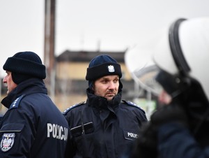 Na pierwszym planie policjant w umundurowaniu służbowym obok niego dwóch policjantów plecami.