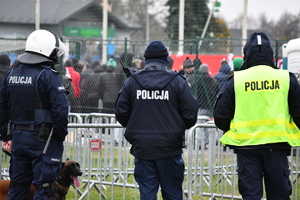 Trzech policjantów plecami, w tle boisko i kibice.