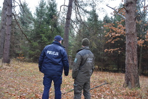 W środku umundurowany policjant i funkcjonariusz straży leśnej idą lasem.