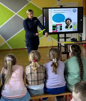 Policjantka w umundurowaniu służbowym trzymająca w dłoni odblaskową opaskę. Dzieci siedzą na ławkach.