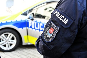 Ramię policjanta w umundurowaniu służbowym z emblematem Posterunku Policji w Cieszanowie, w tle oznakowany radiowóz.