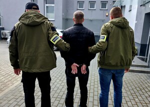 mężczyzna w kajdankach założonych na ręce z tyłu, dwóch policjantów z opaską z napisem "POLICJA".
